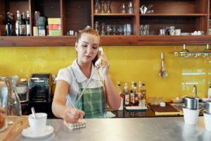 Young woman in apron speaking on phone while standing at counter in coffee shop and writing down order in notepad